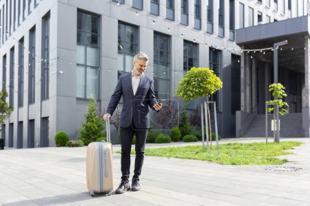 Photo for Mature businessman in suit with suitcase outside office building texting on phone, symbolizing business travel or commute. - Royalty Free Image