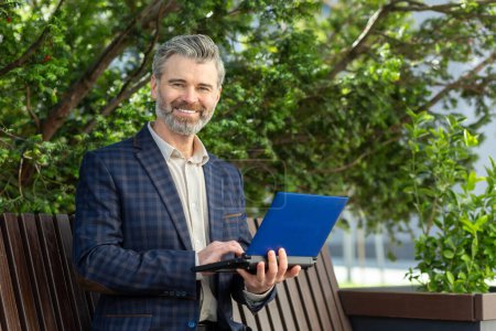 Photo for Cheerful mature male professional in a business suit using a laptop outdoors near an office building, exuding confidence. - Royalty Free Image