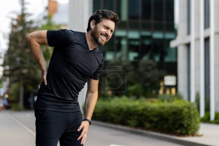 Photo for A young male athlete stands bent over on a city street and grimaces in pain, holding his back. - Royalty Free Image