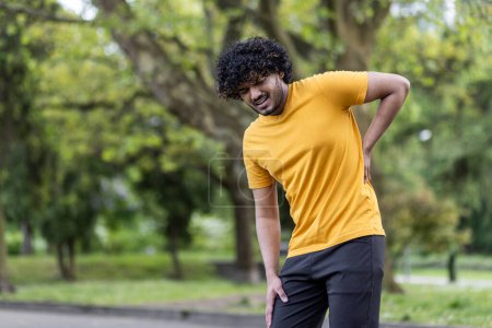 Photo for A young Indian male athlete is standing in the park, bent over and holding his hand behind his back, injured, sprained, feeling terrible pain. - Royalty Free Image