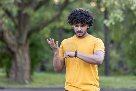 Foto de Joven enfocado con cabello rizado comprobando smartwatch durante el entrenamiento al aire libre en el parque, vistiendo camiseta amarilla casual, verde borroso. - Imagen libre de derechos