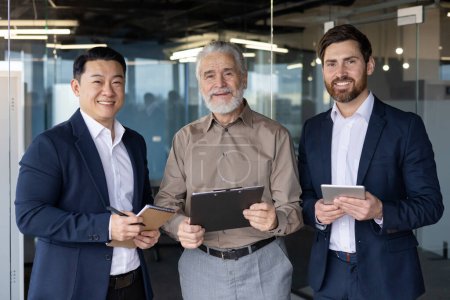 Foto de Tres diversos empresarios profesionales sonriendo y de pie juntos en un entorno de oficina, mostrando el trabajo en equipo y la colaboración. - Imagen libre de derechos