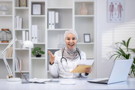 Eine professionelle muslimische Ärztin, die einen Hijab trägt, strahlt Positivität aus, als sie in ihrem hellen Klinikbüro Patienten berät.