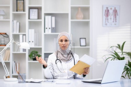Professional woman in hijab and lab coat in a clinic office, engaging in patient consultation with laptop and medical equipment.