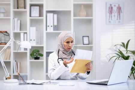 Eine fokussierte muslimische Ärztin in Hijab und weißem Mantel untersucht Dokumente in ihrer Arztpraxis, die die Vielfalt des Gesundheitswesens symbolisieren..