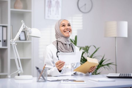 Smiling female Muslim doctor in a hijab and white coat holding papers in a modern clinic office, showcasing professional healthcare.