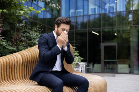 Exhausted mature businessman with a beard sitting outside an office building, covering face with hands in despair.