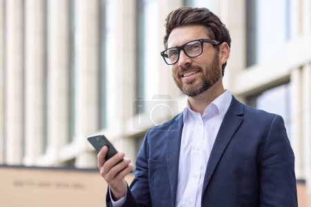 Photo for Mature businessman with glasses and a beard checks his smartphone outside an office building, exuding confidence and professionalism. - Royalty Free Image
