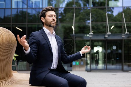 Foto de Empresario barbudo en un traje meditando al aire libre, mostrando el equilibrio entre trabajo y vida por edificios de oficinas. - Imagen libre de derechos