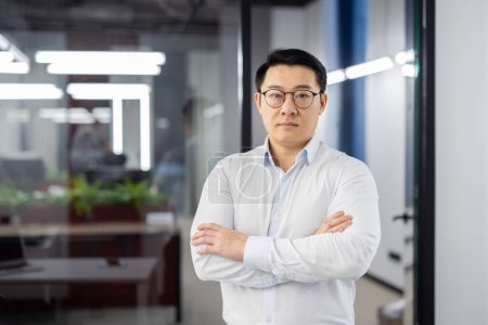 Porträt eines ernsthaften selbstbewussten asiatischen Mannes im Büro am Arbeitsplatz, Geschäftsmann im Hemd mit faltigen Händen und Brille, der in die Kamera blickt, reifer Chef, Finanzbuchhalter.