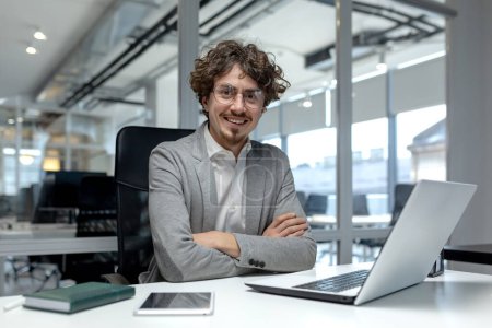 Ein professioneller junger Geschäftsmann mit lockigem Haar, der selbstbewusst an seinem Laptop in einem modernen Büroumfeld arbeitet.