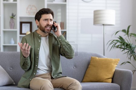 Photo for Adult man with beard appears worried during a phone conversation, seated comfortably in his modern living room. - Royalty Free Image