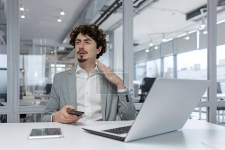 Konzentrierter junger erwachsener Geschäftsmann mit lockigem Haar, der an seinem Schreibtisch in einer modernen Büroumgebung mit Laptop und Smartphone arbeitet.