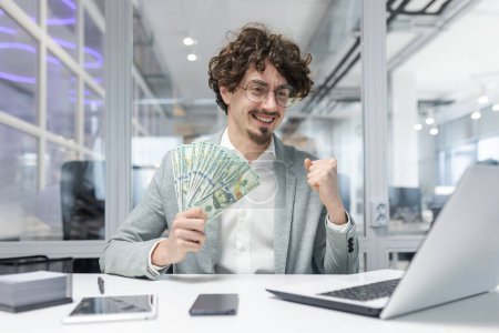 Joyful businessman celebrates financial success at workplace with cash in hand, exuding confidence and achievement in a modern office.