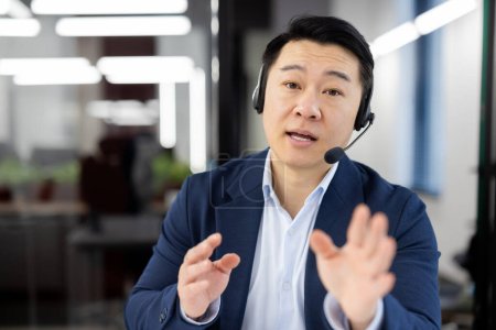 Téléchargez les photos : Agent de soutien masculin asiatique confiant conversant avec des clients utilisant un casque dans un environnement de bureau. - en image libre de droit