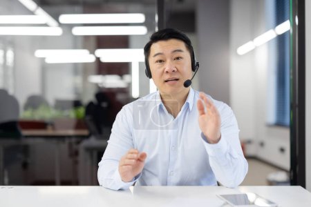 Téléchargez les photos : Homme d'affaires asiatique avec casque dans un bureau moderne fournissant un soutien à la clientèle ou dirigeant une réunion d'équipe, affichant un engagement. - en image libre de droit