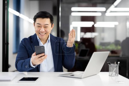 Happy Asian executive engaging in a virtual greeting, waving at the camera while having a video call on his mobile phone.