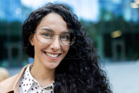 Porträt einer jungen hispanischen Geschäftsfrau mit lockigem Haar und Brille, die während einer Arbeitspause vor einem städtischen Bürogebäude lächelt.