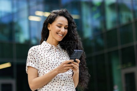 Young, attractive Hispanic businesswoman with curly hair checks her phone with a smile while taking a leisurely walk during her lunch break.