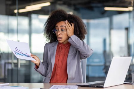 Amazed black woman grabbing head with arm while holding negative statistics in hand and sitting by desktop. Displeased financial worker getting report about inflated expenses and risk of bankruptcy.