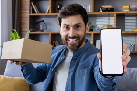 Glücklicher Mann, der ein Paket hält und einen leeren Telefonbildschirm zeigt, ideal für Anzeigen und App-Attrappen, mit einem gemütlichen Zuhause im Hintergrund.
