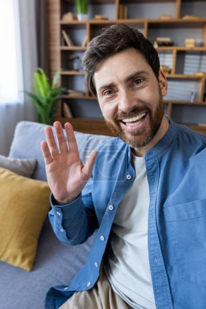 Un hombre alegre y barbudo con ondas ocasionales a la cámara con una sonrisa cálida y acogedora en un ambiente hogareño confortable.