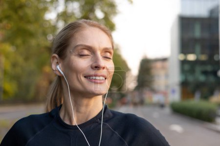 Smiling woman with closed eyes lost in music while using white earphones, enjoying a sunny day in the city.