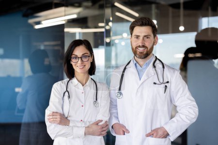 Photo for Two healthcare professionals, a woman and a man in white coats with stethoscopes, smile confidently in a hospital setting. - Royalty Free Image
