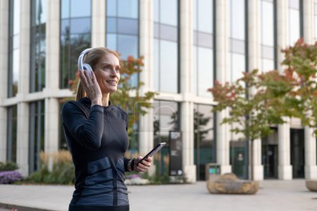Joyful female in sporty outfit using smartphone and headphones outside modern buildings, radiating positivity and active lifestyle.