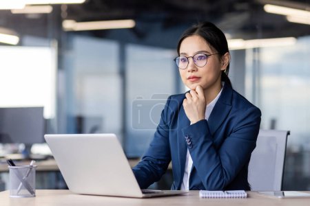 Professional young female executive deep in thought while working at a laptop in a contemporary office setting.