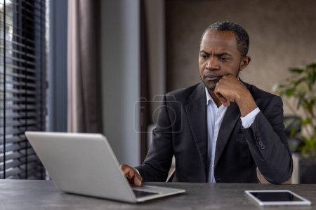 Nachdenkliche männliche Führungskräfte im Anzug vertiefen sich an seinem Schreibtisch im Büro mit Laptop und digitalem Tablet in Betrachtung..