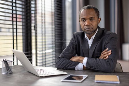 Serious African American businessman in a dark suit sitting with arms crossed at his desk, in a modern office setting.