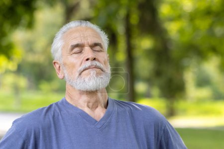 Photo for A gray-haired elderly man relaxes with closed eyes, taking a deep breath outdoors, illustrating peacefulness and wellbeing in nature. - Royalty Free Image