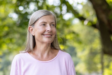 A joyful mature woman with gray hair and a pink top smiles serenely, outdoors with a lush green backdrop.