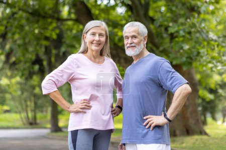 Ein lächelndes älteres Paar in Sportkleidung posiert selbstbewusst während einer Fitnesseinheit in einem ruhigen Park mit grünen Bäumen.