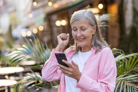 Femme âgée dehors, poing pompé dans la victoire, souriant à son smartphone, capturant la joie des bonnes nouvelles.