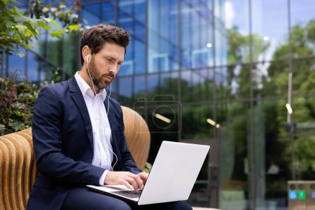 Téléchargez les photos : Entrepreneur professionnel masculin avec écouteurs utilisant un ordinateur portable à l'extérieur de l'immeuble de bureaux moderne, se concentrant sur le travail. - en image libre de droit