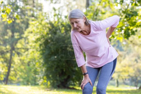 Aktive Seniorin in Sportkleidung, die beim Sport in einem sonnenbeschienenen Park unangenehm ihr Knie berührt, stellt gesundheitliche Probleme dar.