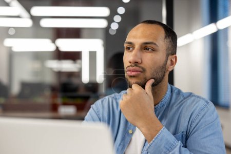 Photo for A contemplative young professional in a well-lit office environment, caught in a moment of thoughtfulness while gazing away from his laptop screen. His expression conveys concentration and deep - Royalty Free Image