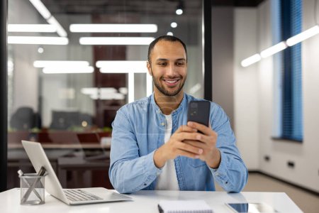 Ein junger männlicher Profi in einem lässigen blauen Jeanshemd unterbricht die Arbeit, um auf sein Handy zu schauen. Das moderne Büroumfeld bietet ein entspanntes, aber produktives Ambiente.