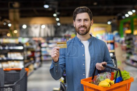 Un joven adulto con una tarjeta de crédito y una cesta llena de productos frescos, listo para pagar en un supermercado moderno. Destacando la conveniencia y el estilo de vida.