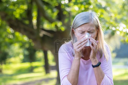 Nahaufnahme einer älteren grauhaarigen Frau, die draußen in einem Park steht und sich mit einer Serviette die Nase abwischt, unter einer laufenden Nase und saisonalen Allergien leidet.