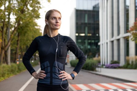 A fitness-focused woman pauses during her run in the city, displaying determination and confidence. Wearing sportswear and earphones, she embodies an active lifestyle against an urban backdrop.