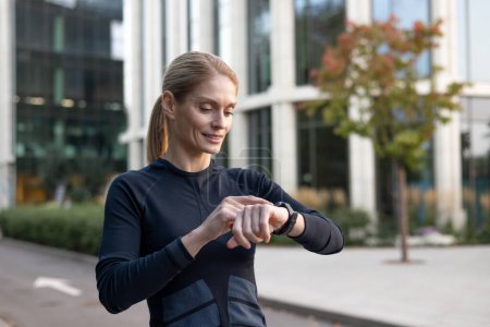 A focused female athlete pauses during her urban workout to monitor her performance on a smartwatch. Capturing a moment of technology integrating with fitness in a city environment.