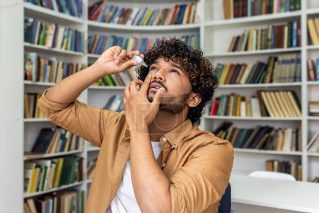A young adult male is captured applying eye drops while standing in a cozy home library. The image reflects self-care and health management against a backdrop of bookshelves filled with books.
