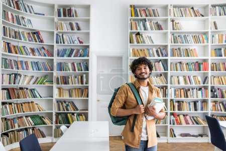 Photo for A cheerful young adult with curly hair and a backpack standing in a bright library, holding several books, exuding a vibe of learning and positivity amid shelves full of books. - Royalty Free Image