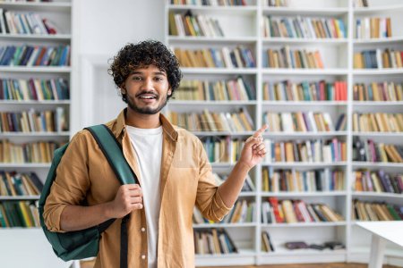 Ein fröhlicher junger Mann mit lockigem Haar und lässigem Outfit steht in einer Bibliothek. Auf einer Schulter trägt er einen Rucksack und zeigt mit einem Lächeln auf etwas aus dem Rahmen.