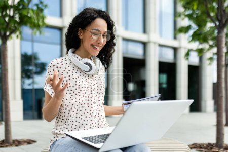 A young female professional engages in a video call while working on her laptop outside a modern office building, portraying flexibility and modern work lifestyle.