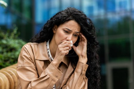 Una mujer de negocios angustiada con atuendo profesional está sentada afuera, limpiando lágrimas con un pañuelo, una mirada de tristeza y preocupación en su cara en medio de un telón de fondo urbano.