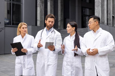 Group of four healthcare workers in lab coats engaged in discussion, holding tablets and documents, outside modern clinic.